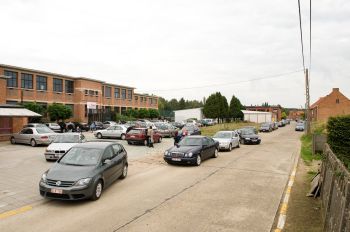 Foto van voorkant van schoolgebouw met daarvoor een groot terrein waarop wagens kriskras geparkeerd staan. Ook op de weg ervoor staat een hele rij wagens geparkeerd.
